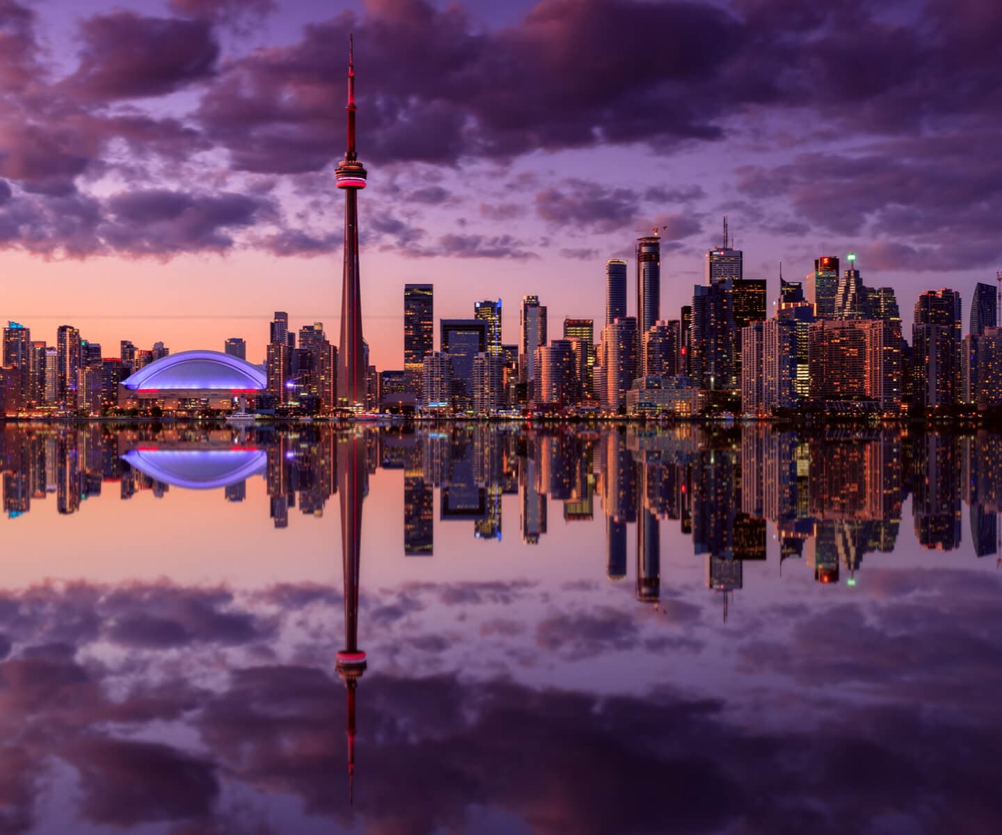 Toronto skyline at dusk, featuring the CN Tower and Rogers Centre, reflects on calm water
