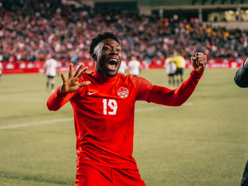 Soccer player in a red Canada jersey celebrates on the field with raised hands in a packed stadium