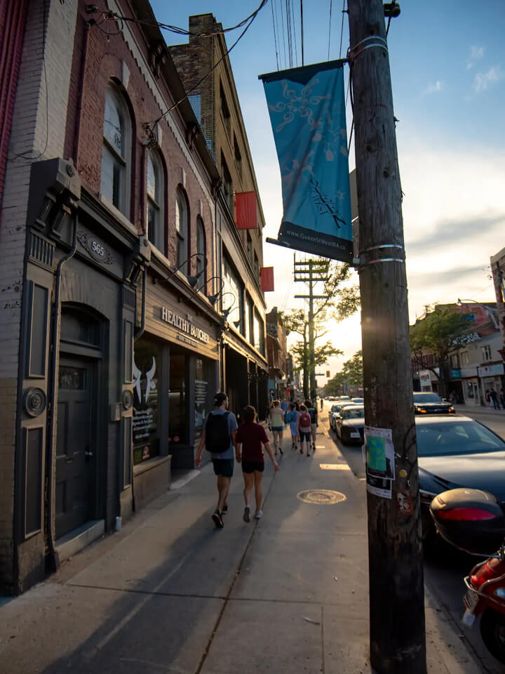 People walking on a busy sidewalk on Queen St West at sunset with glowing neon signs